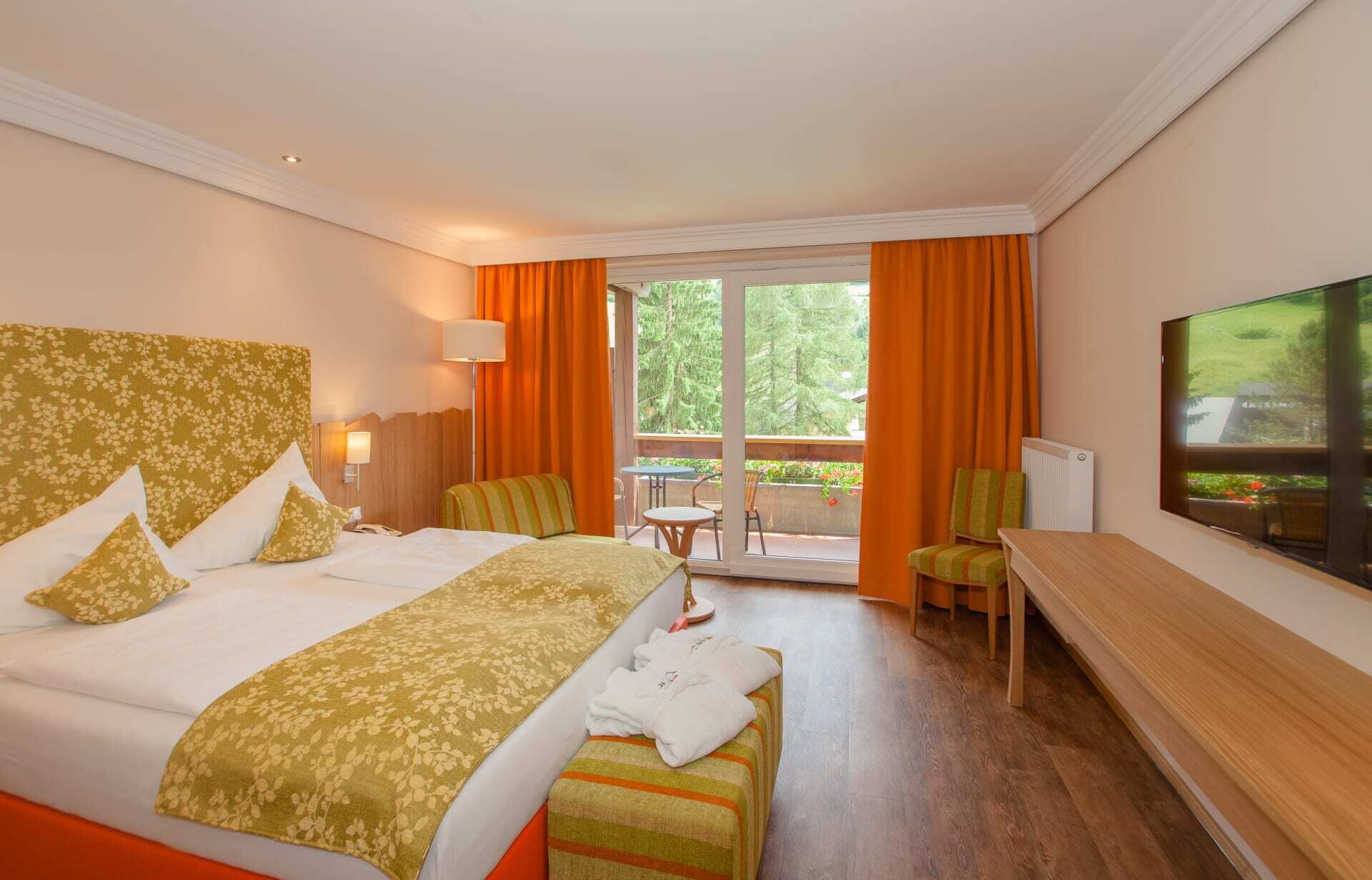 Helles Hotelzimmer mit Doppelbett, Sitzecke und großen Fenstern mit orangefarbenen Vorhängen