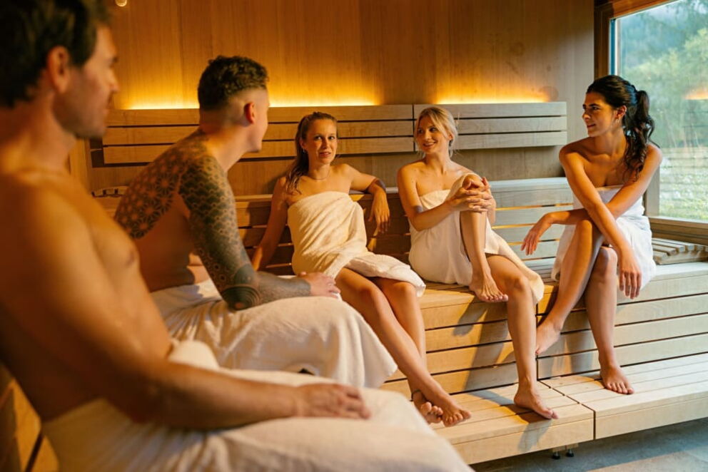 Fünf Personen sitzen in einer Sauna, umhüllt von Handtüchern und in entspannter Konversation.