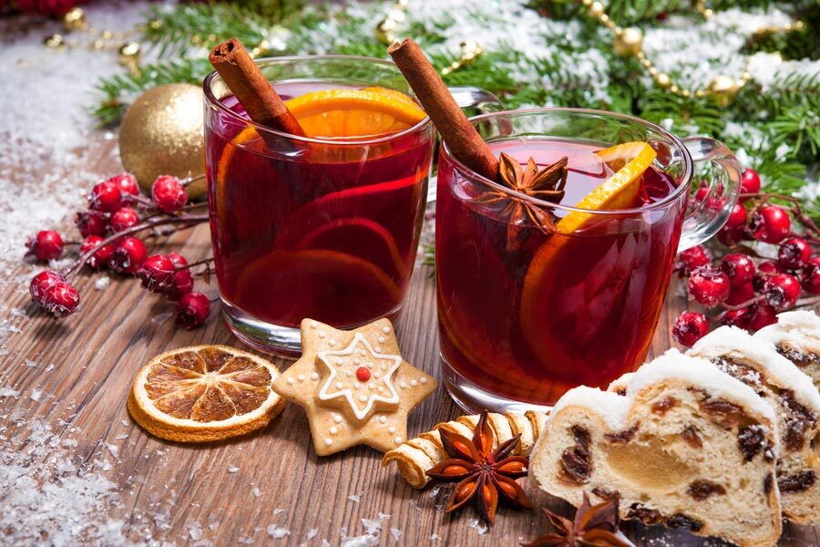 Zwei Gläser Glühwein mit Orangenscheiben und Zimtstangen auf einem Holzuntergrund, umgeben von Keksen und Stollen.