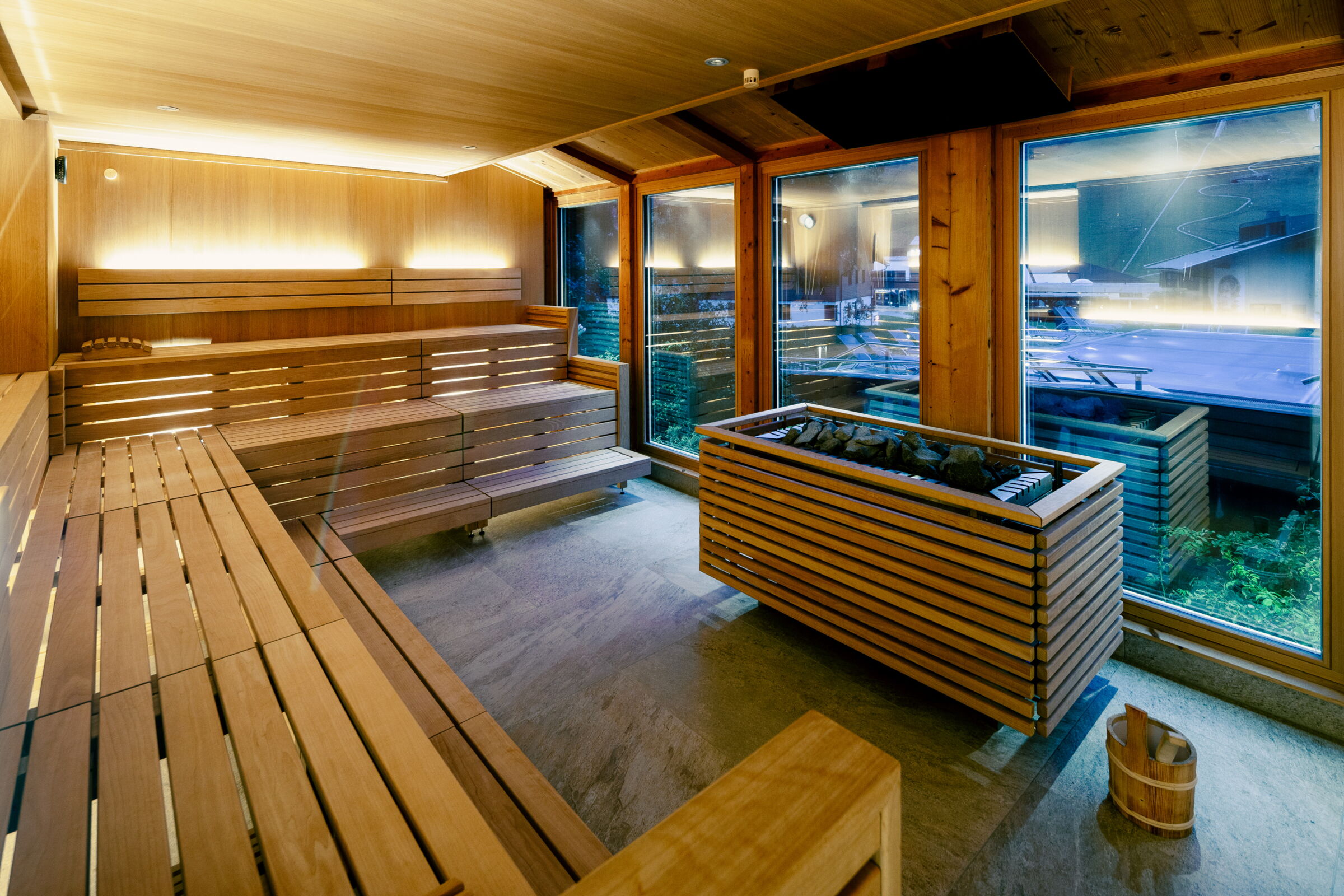 Innenansicht einer modernen Sauna mit Holzliegen und großen Fenstern, Steinofen.