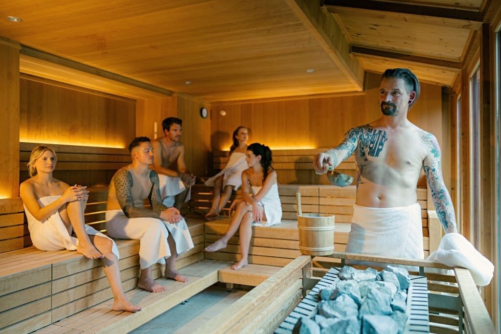 Fünf Personen in einer Holzsauna, die sich entspannen und unterhalten. Eine Person steht und gestikuliert.