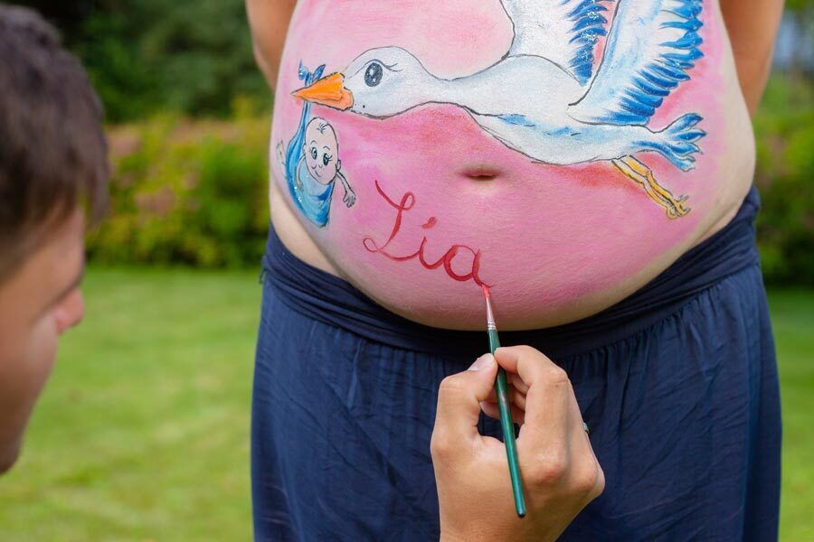 Un uomo che dipinge una pancia incinta, dipinge una cicogna che porta un bambino.