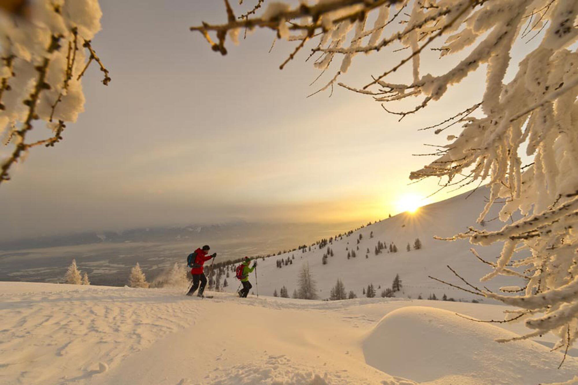 Zwei Wanderer durchqueren eine schneebedeckte Landschaft bei Sonnenuntergang, umgeben von Bäumen und Bergen.