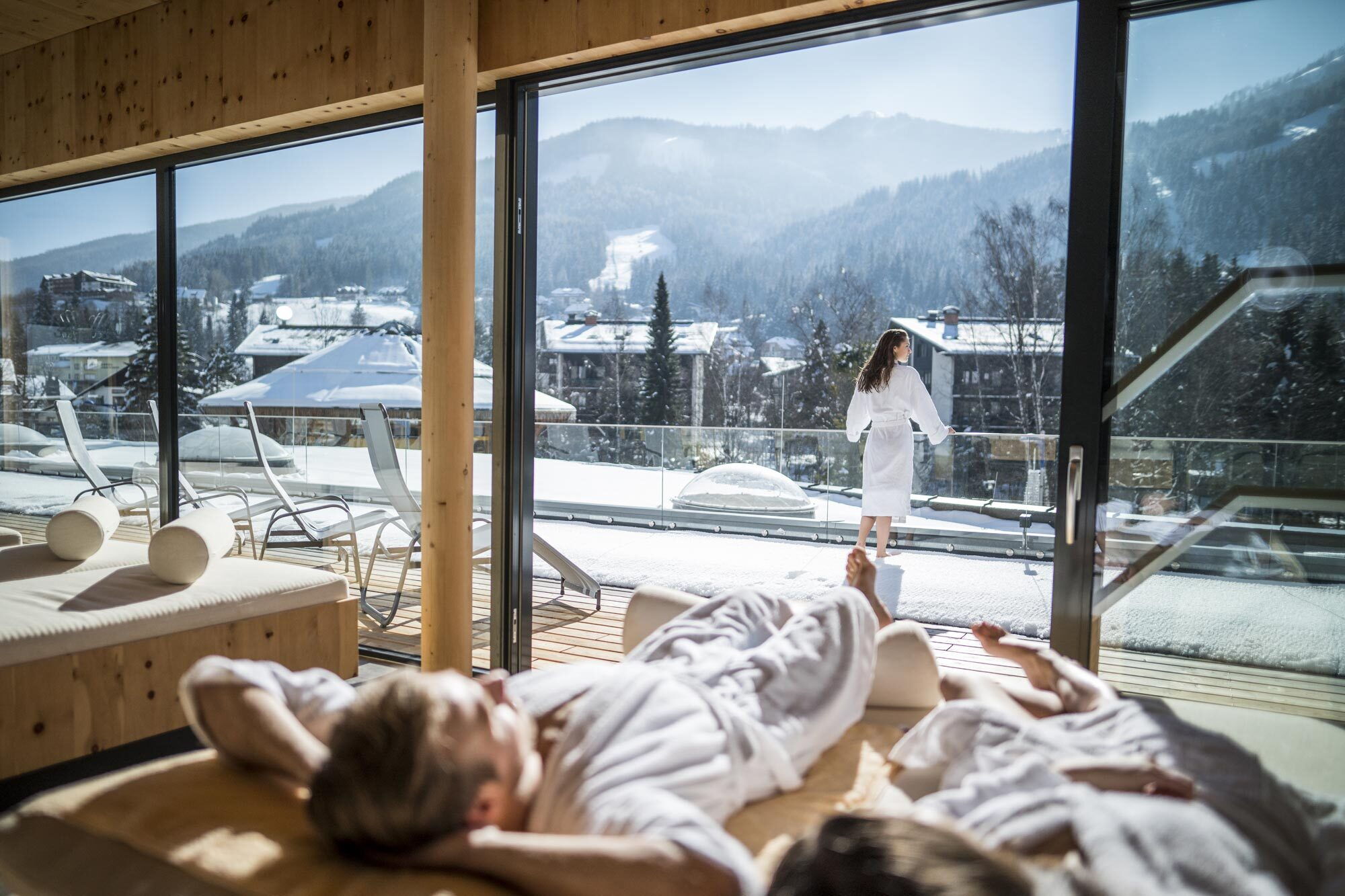 Ruhige Spa-Landschaft mit zwei Personen in Bademänteln auf einer Liege und Blick auf schneebedeckte Berge.