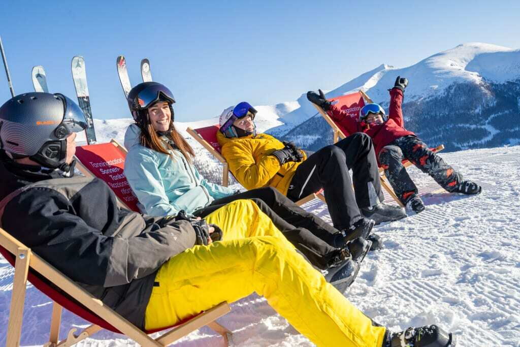 Vier Personen entspannen in Liegestühlen auf einem schneebedeckten Hang, Skier im Hintergrund, blauer Himmel