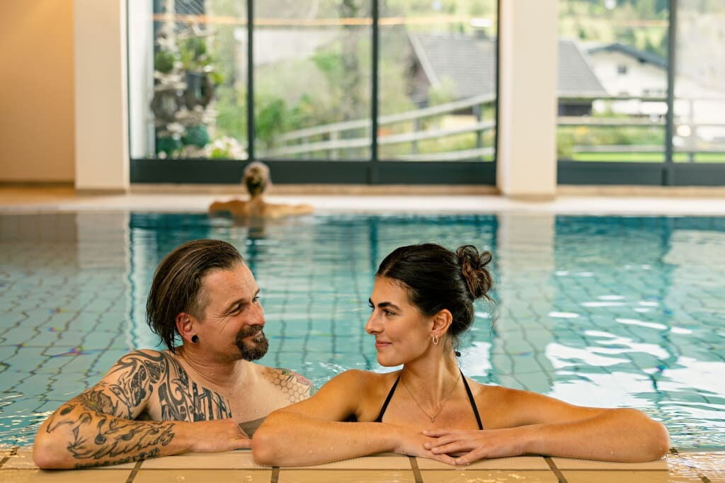 Ein Paar entspannt zusammen in einem Swimmingpool mit natürlichem Hintergrund und einer Person, die im Wasser schwimmt.