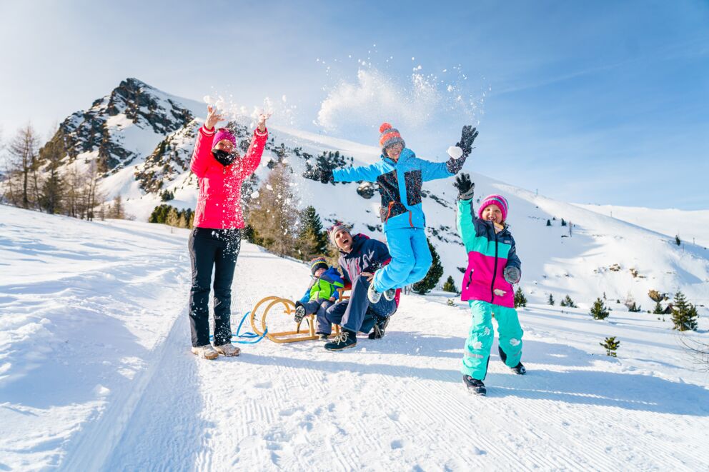Vier Personen in einer verschneiten Landschaft, zwei Kinder spielen und ein Erwachsener fährt Schlitten.