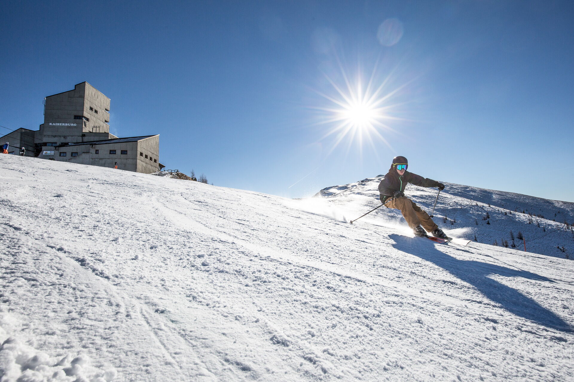 Skifahrer auf einer schneebedeckten Piste mit Bergresort im Hintergrund und klarem Himmel