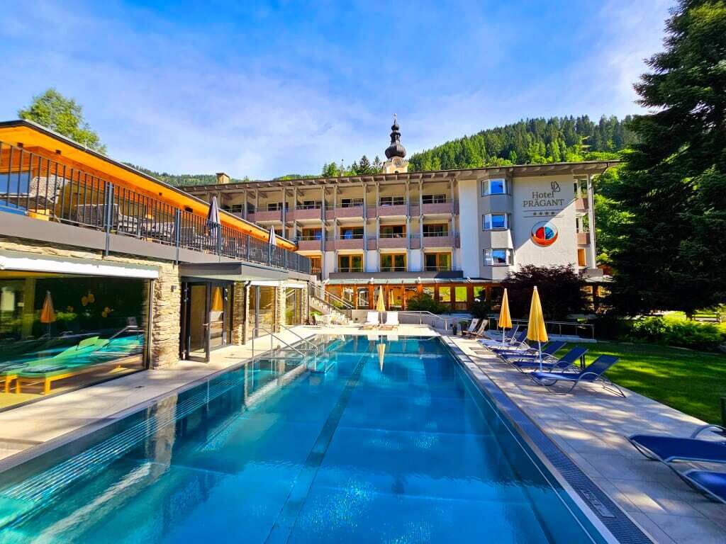 Hotel mit Schwimmbad vor einer landschaftlich gestalteten Außenanlage und Bergen