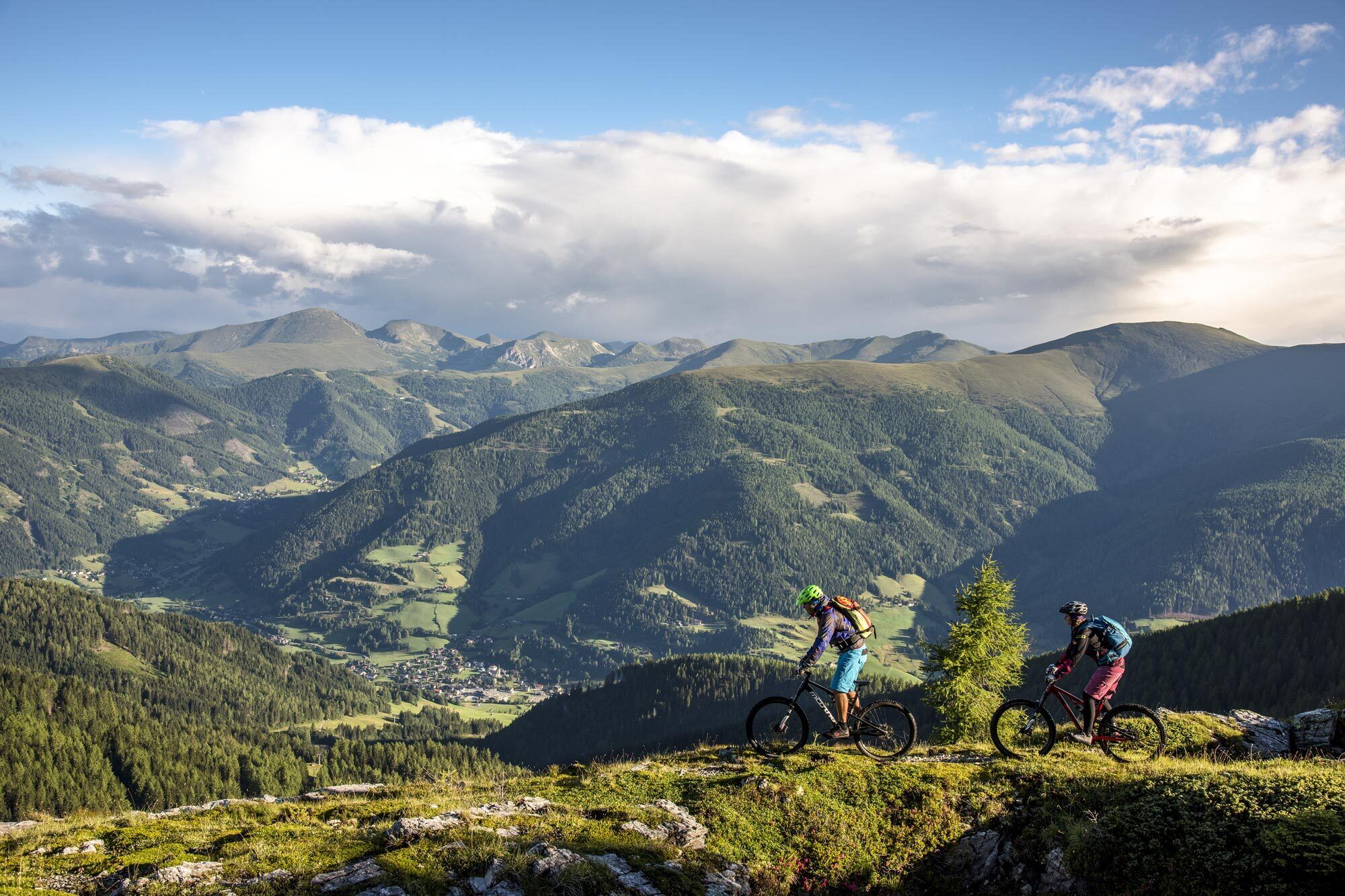 Zwei Mountainbiker fahren einen Pfad entlang, umgeben von Bergen und grünen Tälern.
