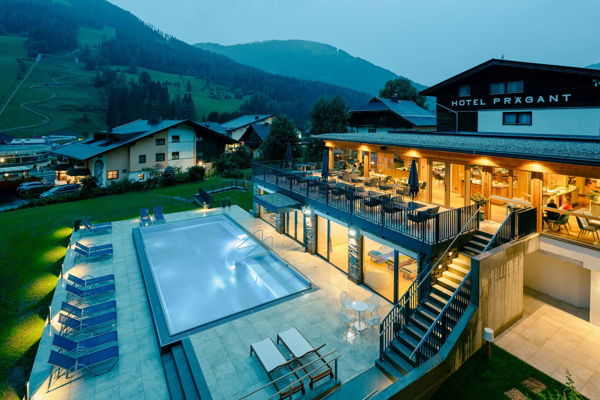 Außenansicht eines Hotels mit modernem Design, Swimmingpool und Sitzgelegenheiten vor Berglandschaft bei Abendlicht.