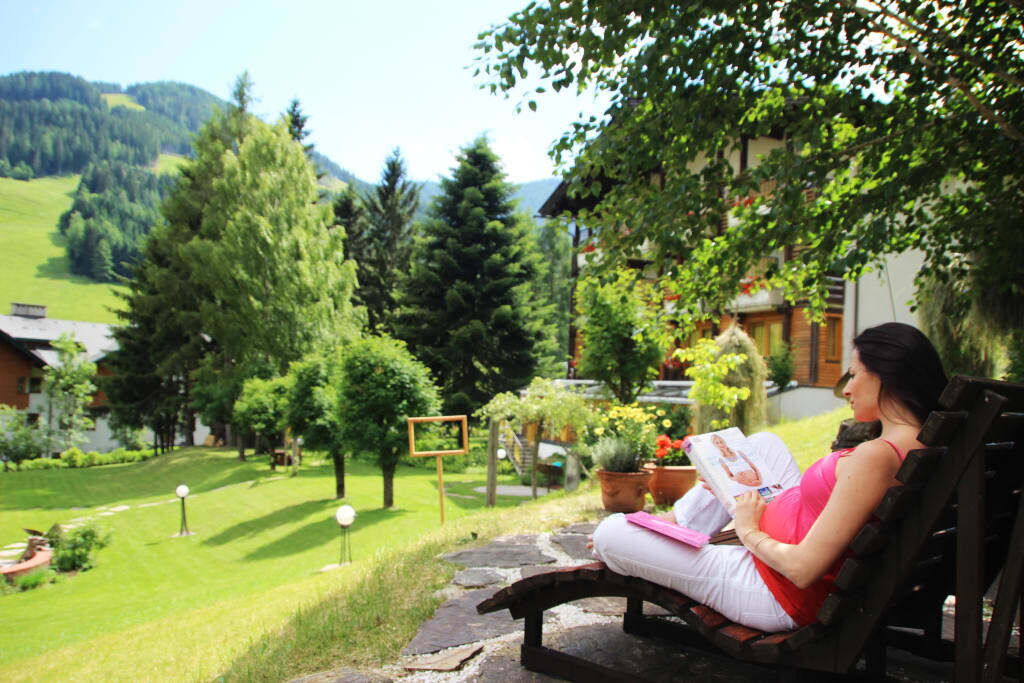 Eine Frau sitzt auf einer Liege im Freien und liest eine Zeitschrift, umgeben von Bäumen und Bergen.