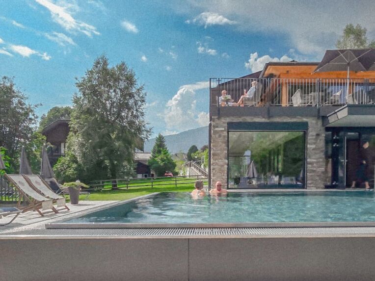 Außenpool in einer Hotelanlage umgeben von Bäumen und Bergen im Hintergrund mit Sonnenliegen