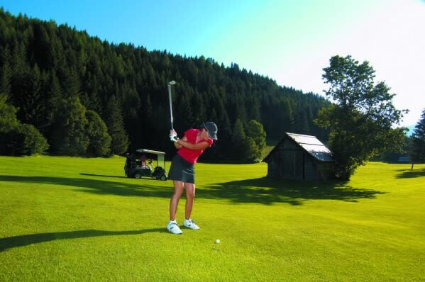 Golfer führt einen Schwung auf einem Golfplatz aus, im Hintergrund Wald und ein Golfcart.