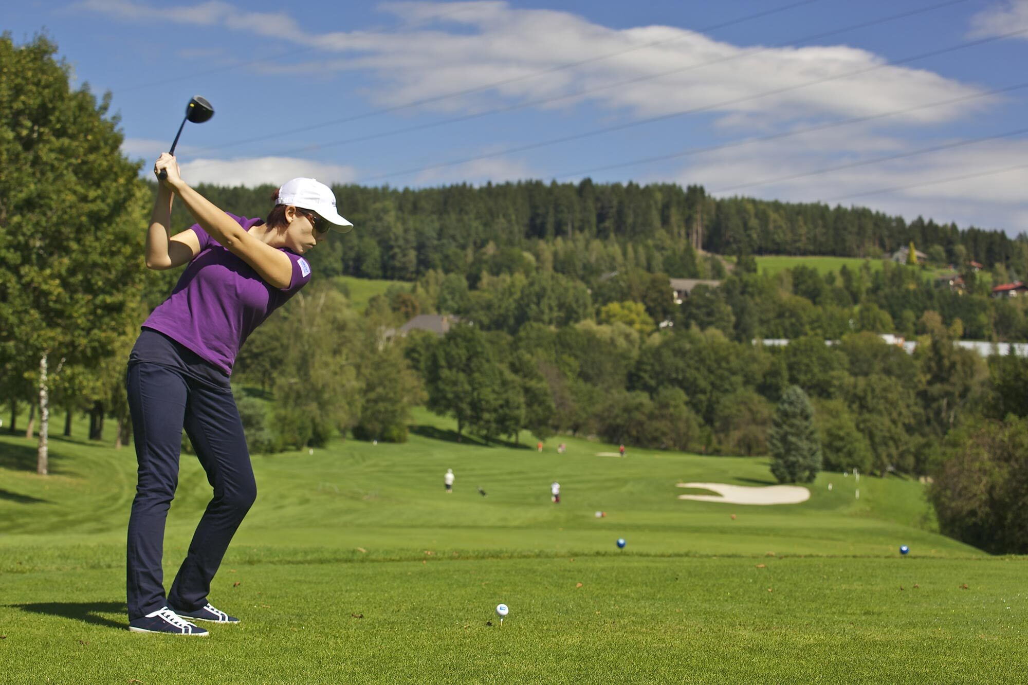 Frau beim Golfspielen auf einem gepflegten Platz umgeben von Bäumen und Himmel mit Sicht auf den Golfplatz