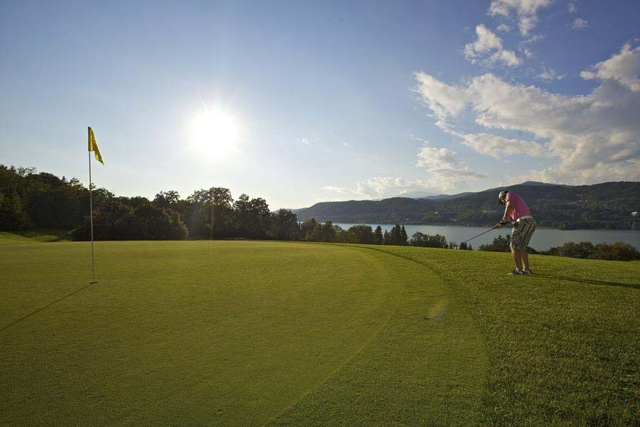 Golfer puttet auf einem gepflegten Golfplatz mit Blick auf Hügel und einen See unter klarem Himmel.