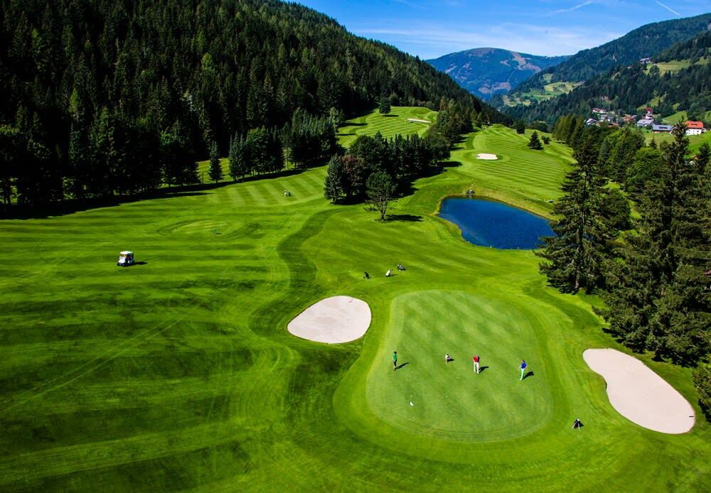 Landschaft mit Golfplatz, fairways, Sandbunkern und einem Teich umgeben von Bäumen und Bergen