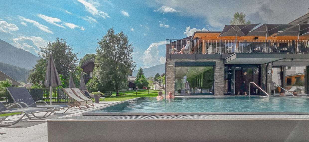Außenpool in einer Hotelanlage umgeben von Bäumen und Bergen im Hintergrund mit Sonnenliegen