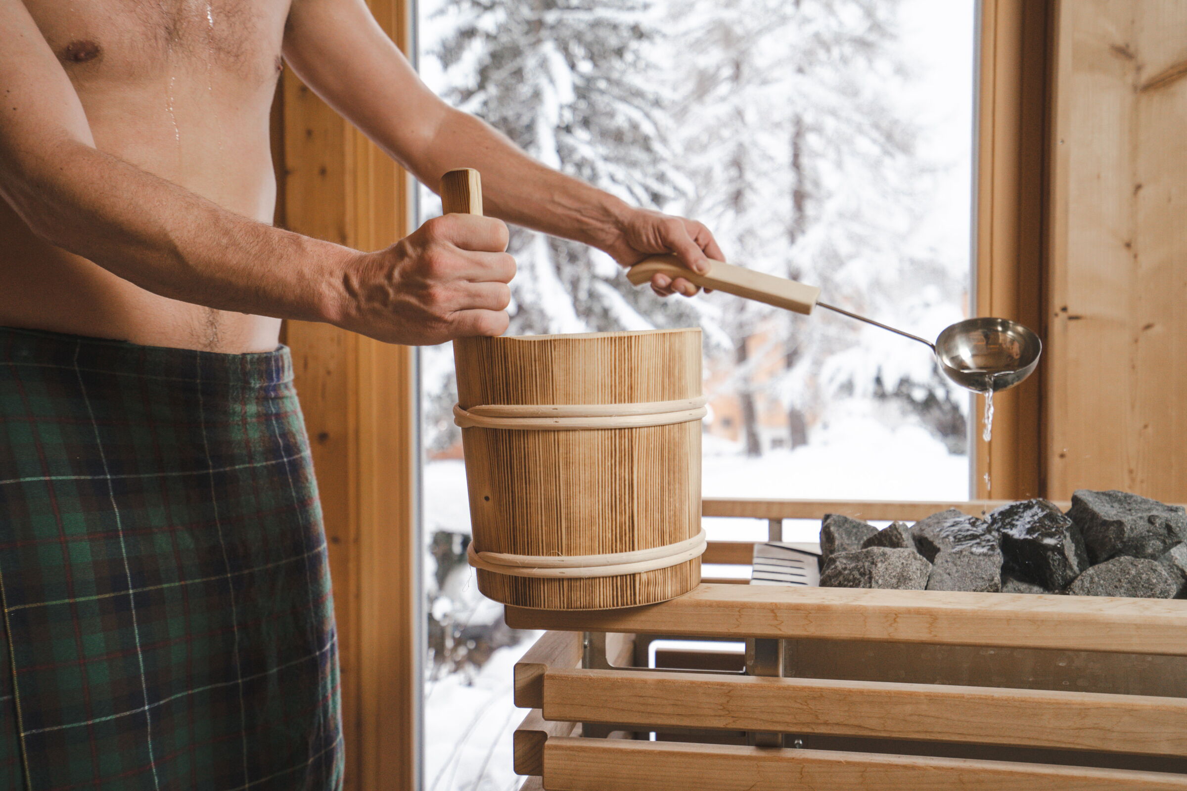 Person bereitet Aufguss in der Sauna vor, während Schnee draußen sichtbar ist, entspannende Atmosphäre