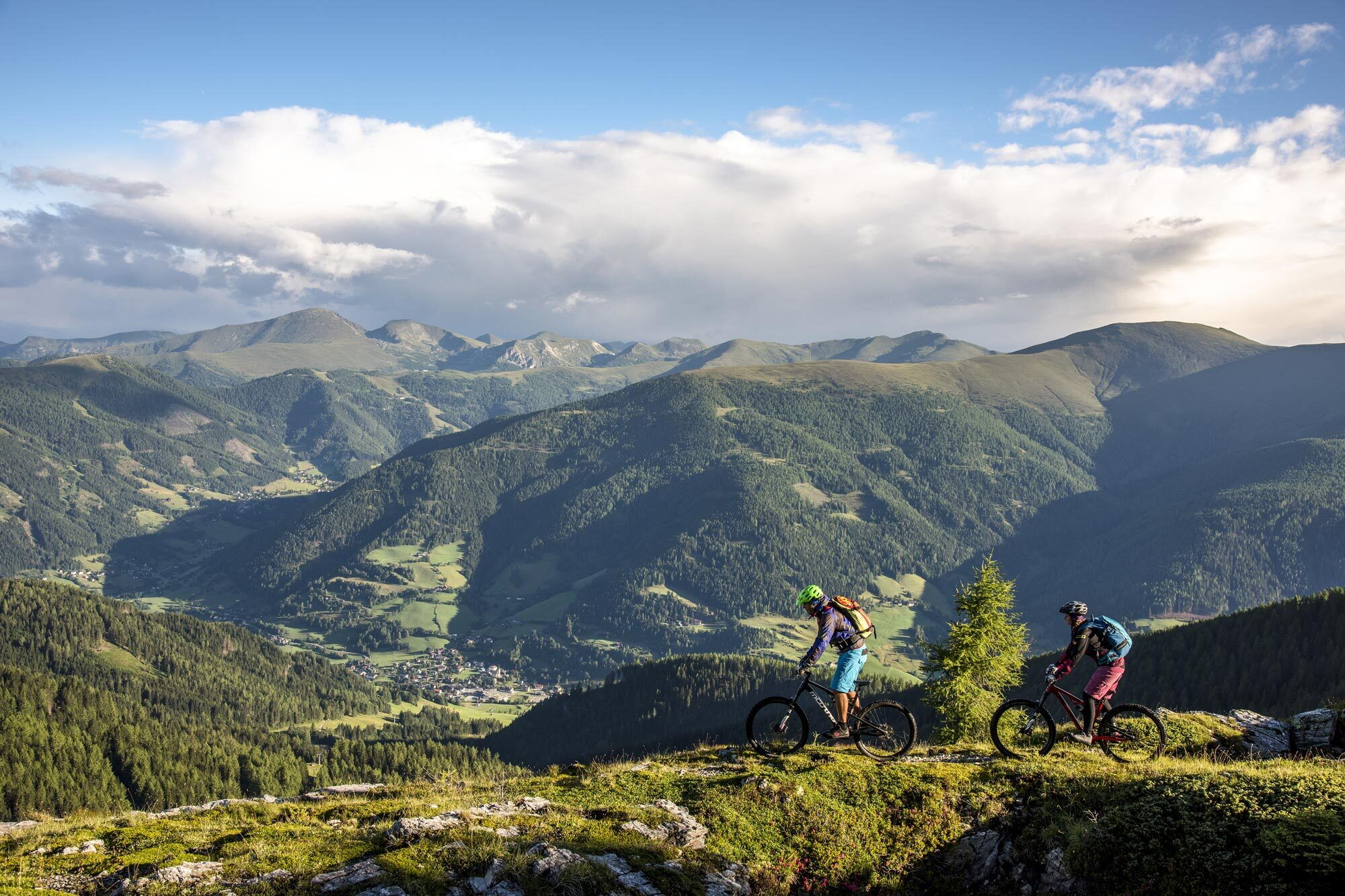 Zwei Mountainbiker fahren einen Pfad entlang, umgeben von Bergen und grünen Tälern.
