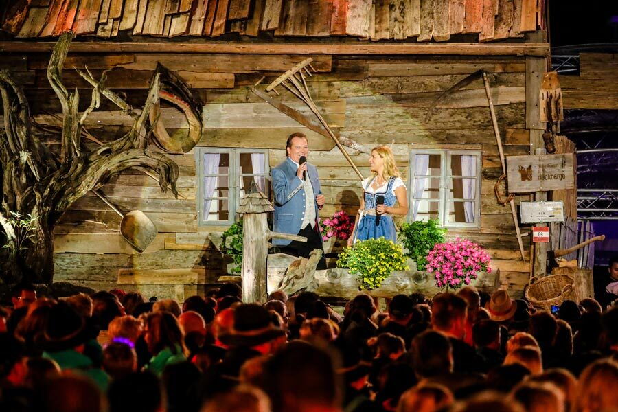 Two presenters on an open air stage with an Austrian mountain hut backdrop