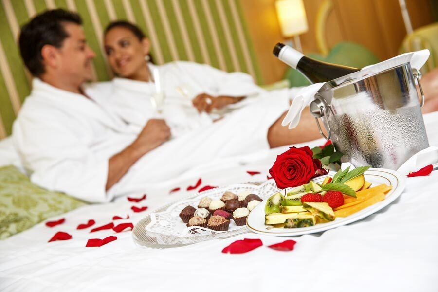 Paar in Bademänteln sitzt auf dem Bett mit Champagner und Schokoladenteller umgeben von Rosenblättern.