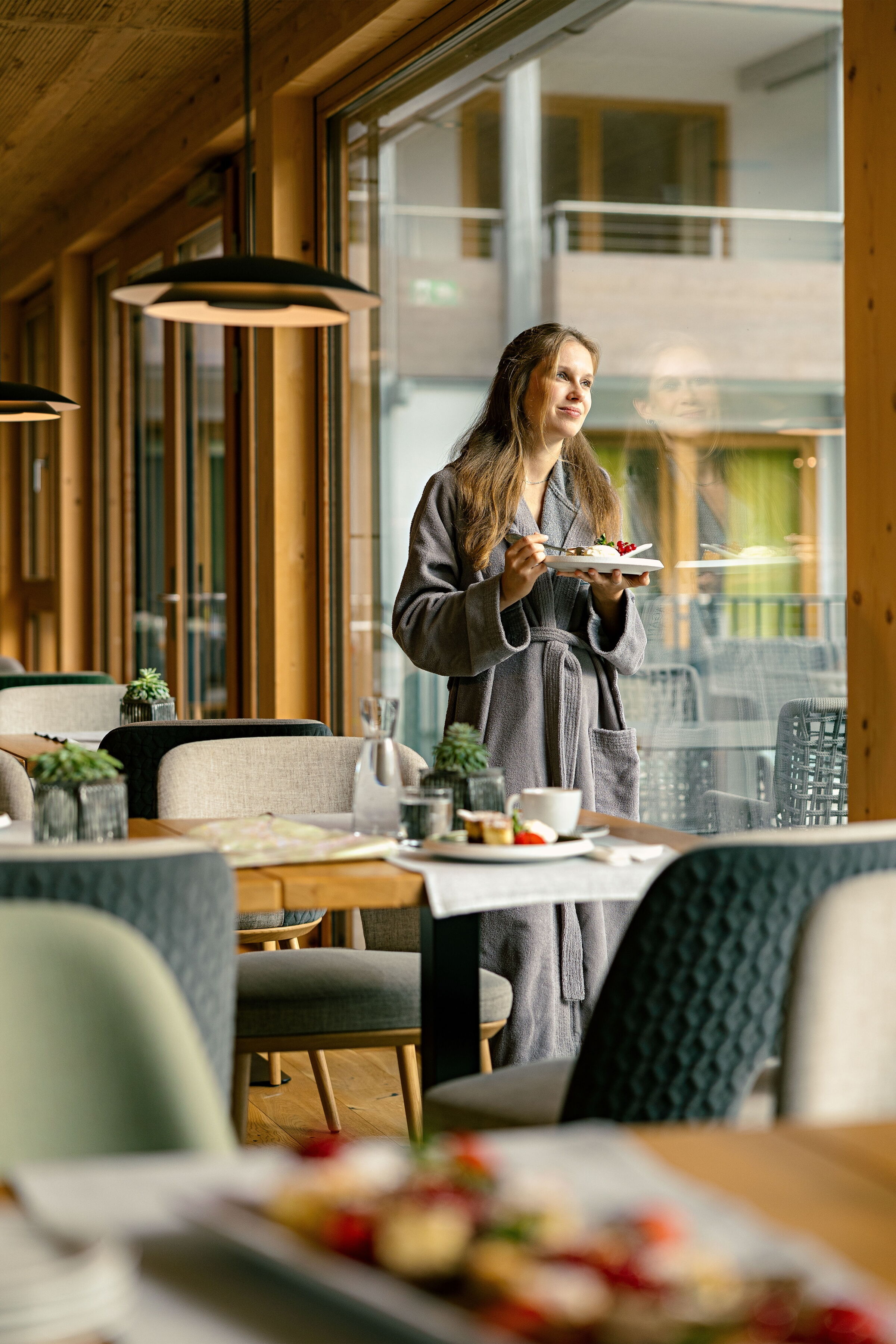 Frau in Bademantel genießt gesunde Speisen im Spa-Restaurant mit Ausblick auf die Natur.