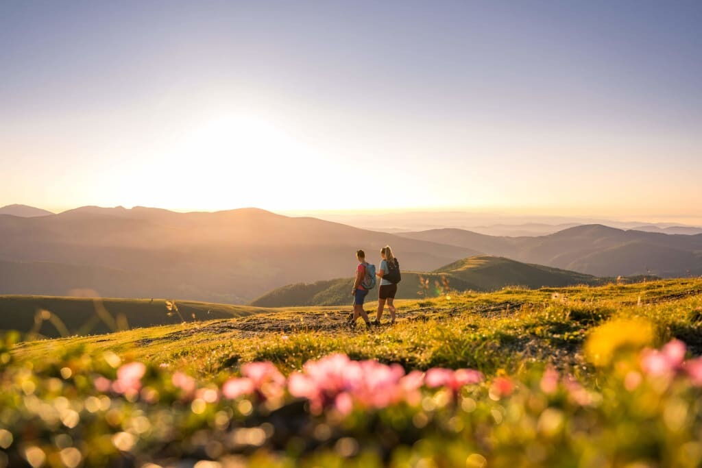 Zwei Personen wandern auf einem grasbewachsenen Hügel bei Sonnenuntergang, Berge im Hintergrund