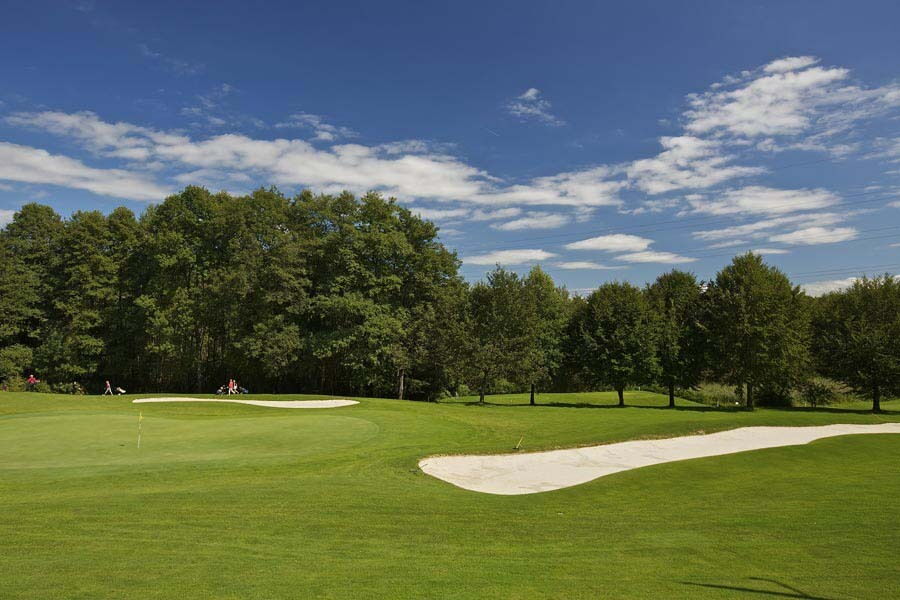 Szenische Golfplatzlandschaft mit grünen Fairways und Sandbunkern umgeben von Bäumen und Himmel.