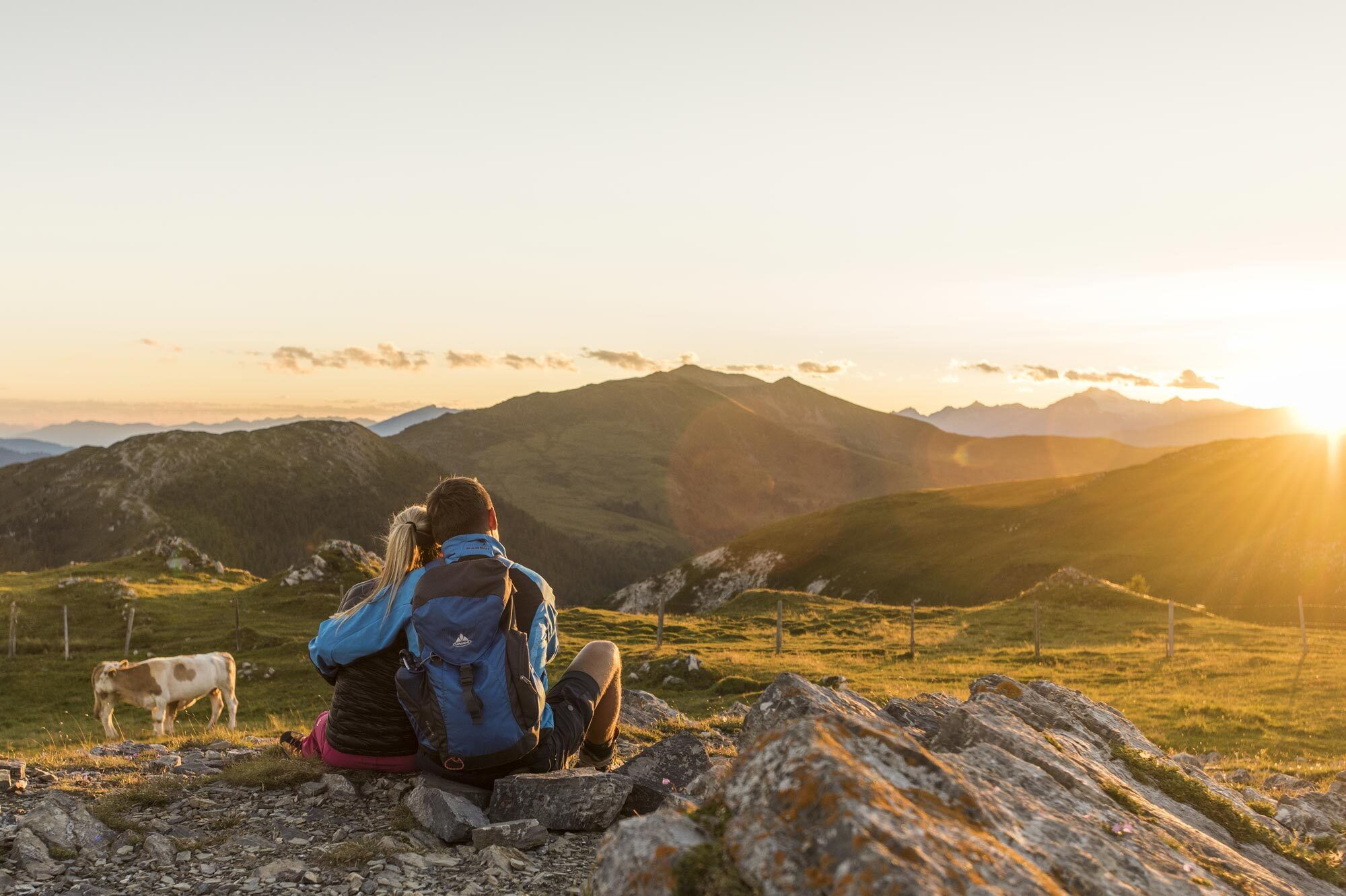 Paar sitzt auf Felsen und blickt auf eine Berglandschaft bei Sonnenuntergang mit weidenden Kühen.