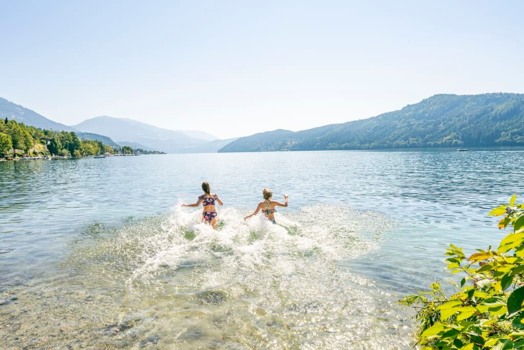Zwei Kinder laufen ins Wasser eines Sees und spritzen dabei, umgeben von einer sonnigen Atmosphäre