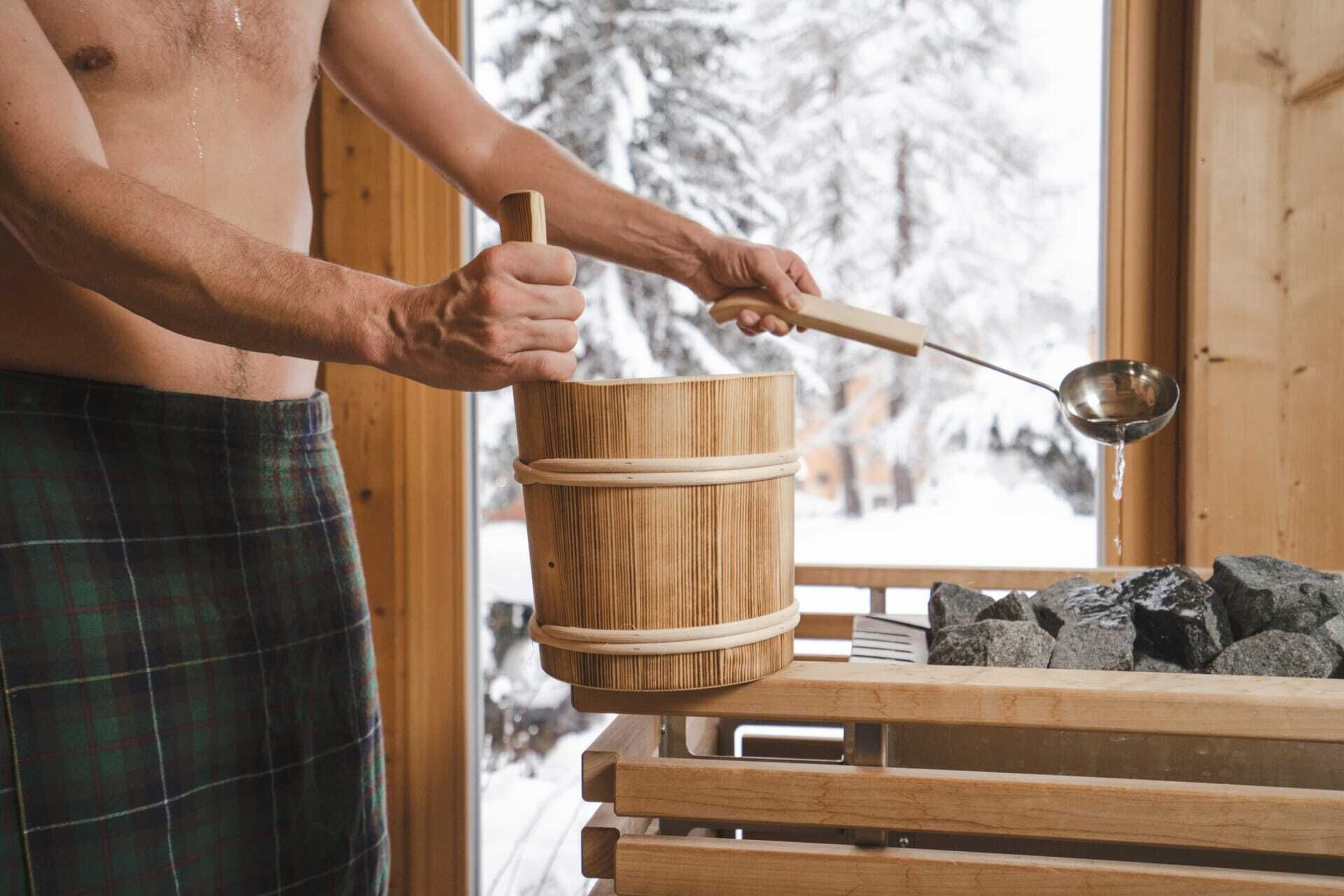 Person mit Holzschöpfkelle bereitet Wasseraufguss auf heißen Steinen in einer Sauna vor