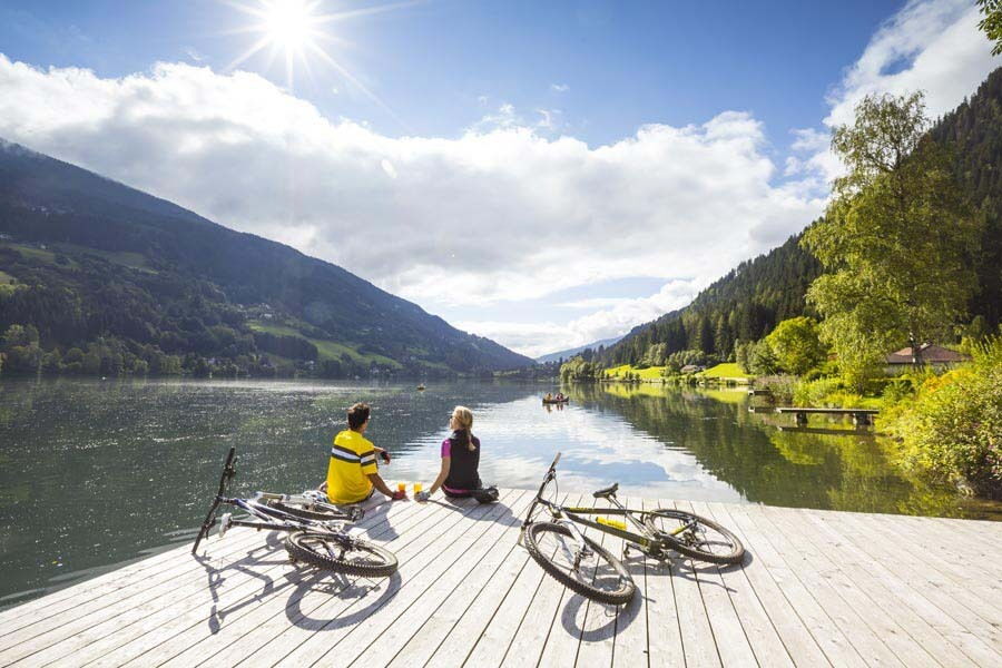 Paar sitzt an einem Holzsteg am See, Fahrräder in der Nähe, umgeben von Bergen und Grün.