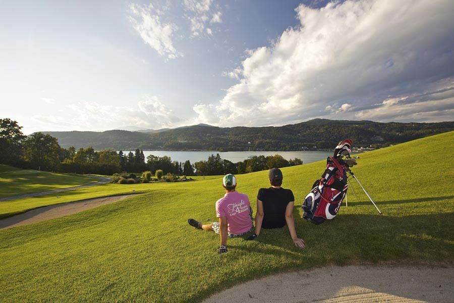 Zwei Personen entspannen auf einer Wiese mit Blick auf einen See und Berge, Golfbag daneben.