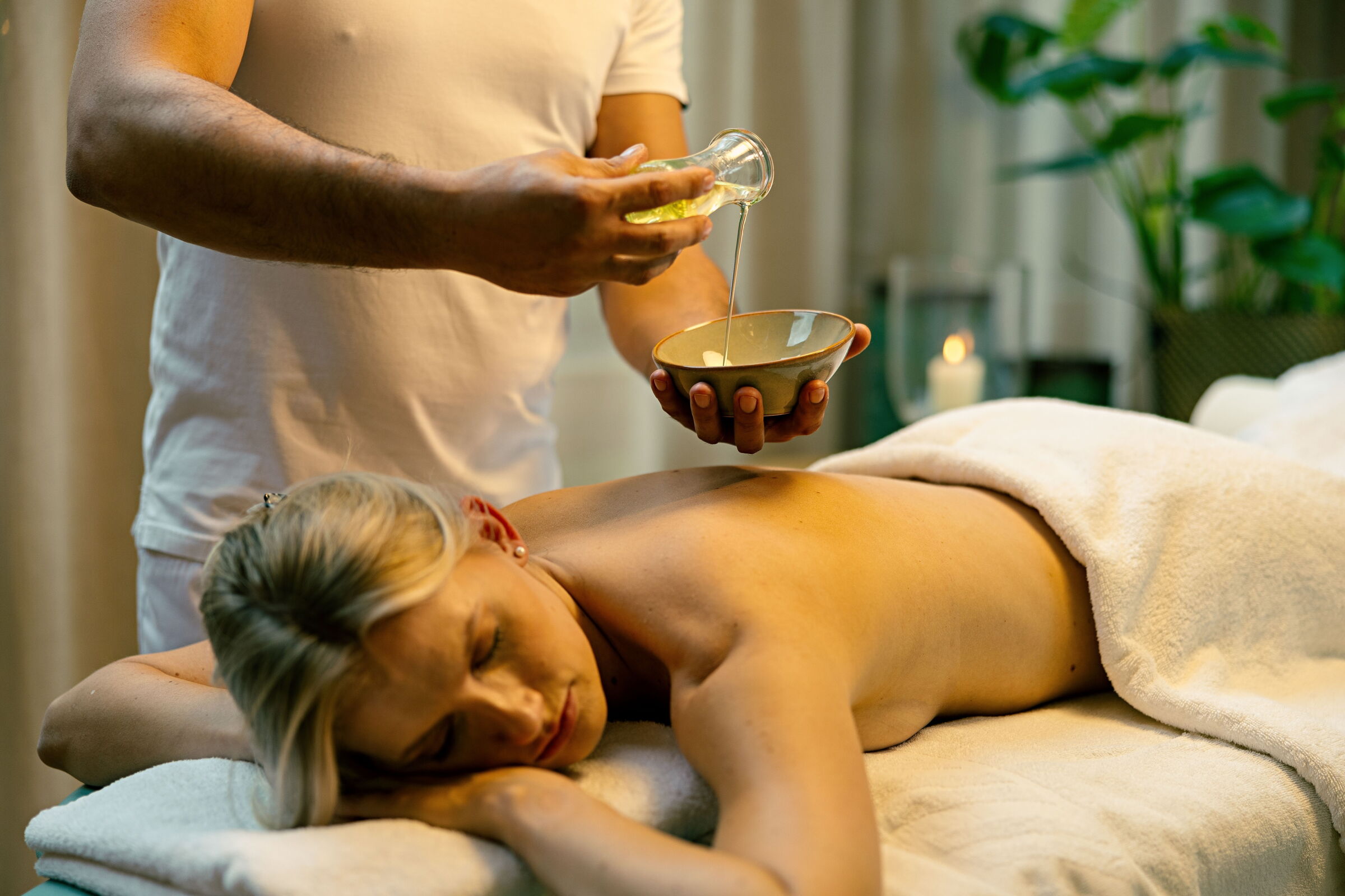 Therapeut gießt ätherische Öle in eine Schüssel neben einer liegenden Frau auf der Massageliege.