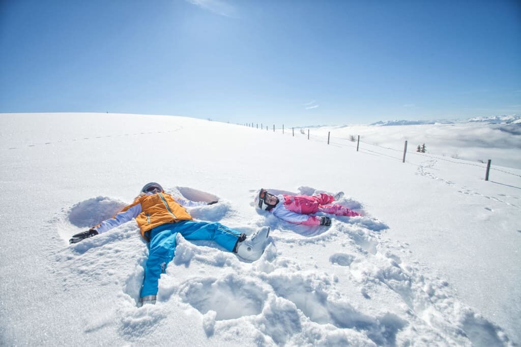Zwei Kinder liegen im Schnee und formen Schneegestalten an einem sonnigen Wintertag.
