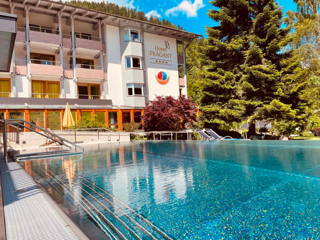 Außenansicht eines Hotels mit Schwimmbad, umgeben von Bäumen und Bergen im Hintergrund.