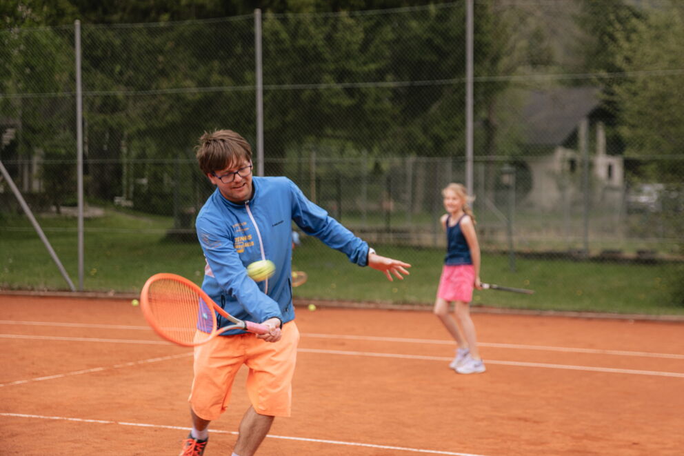 Mann spielt Tennis auf einem Platz mit einem Mädchen im Hintergrund, das sich auf einen Schlag vorbereitet.