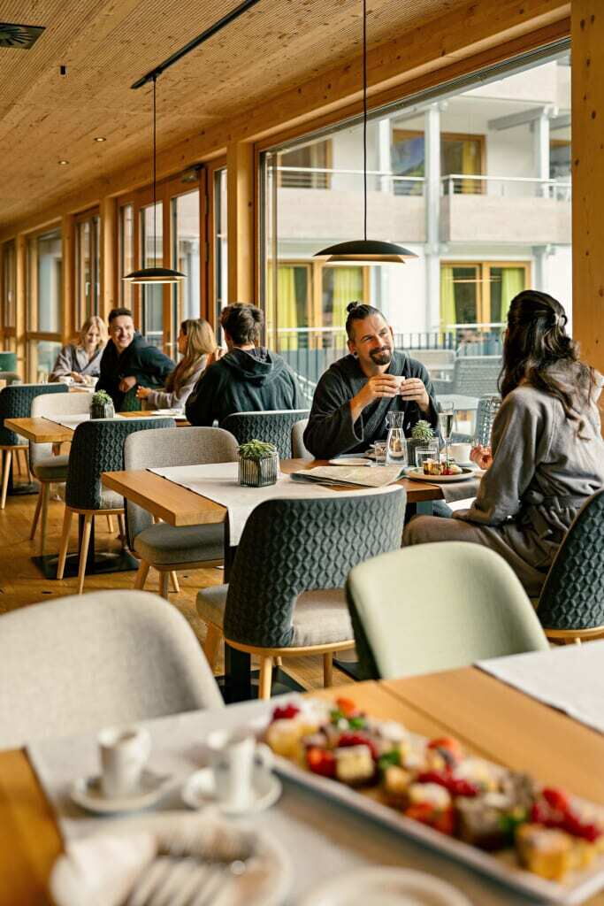 Innenansicht eines modernen Restaurants mit Gästen an Tischen im Gespräch und großen Fenstern.