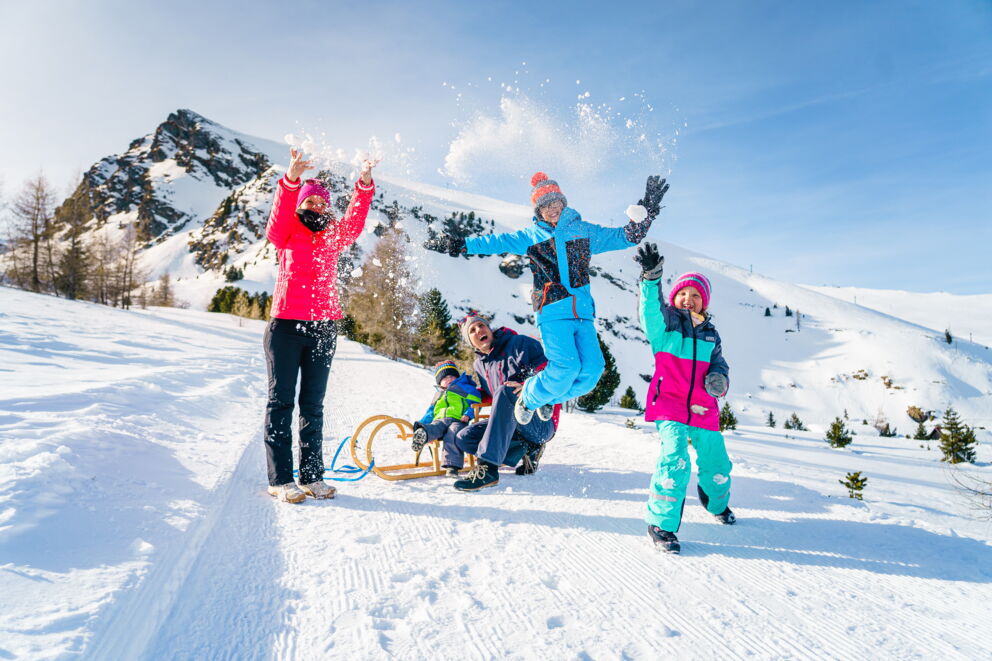 Vier Personen in einer verschneiten Landschaft, zwei Kinder spielen und ein Erwachsener fährt Schlitten.