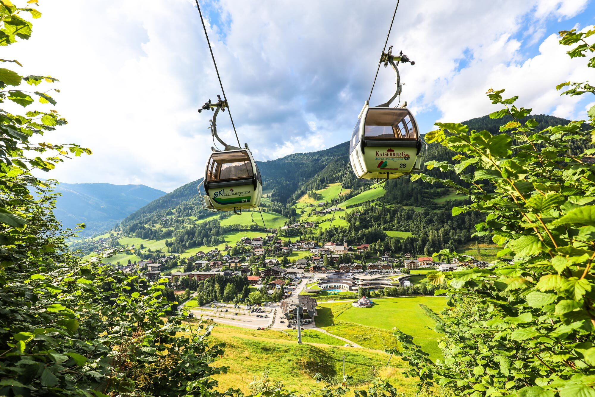 Zwei Seilbahnen schweben über eine grüne Landschaft mit Bergen im Hintergrund und einem Dorf.