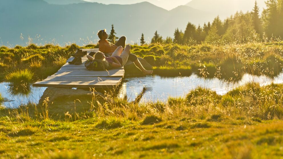 Two hikers lying on a small wooden bridge by a pond