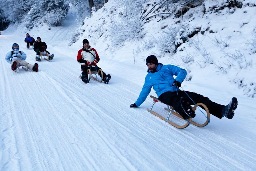 Gruppe von Menschen rodeln fröhlich einen schneebedeckten Hang hinunter in einer Winterlandschaft.