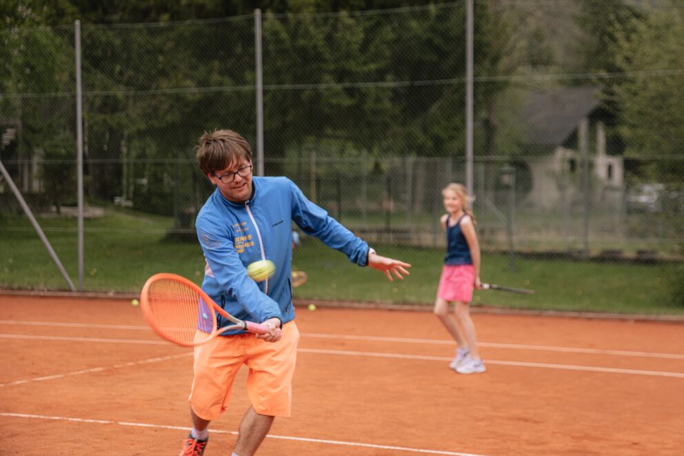 Mann spielt Tennis auf einem Platz mit einem Mädchen im Hintergrund, das sich auf einen Schlag vorbereitet.