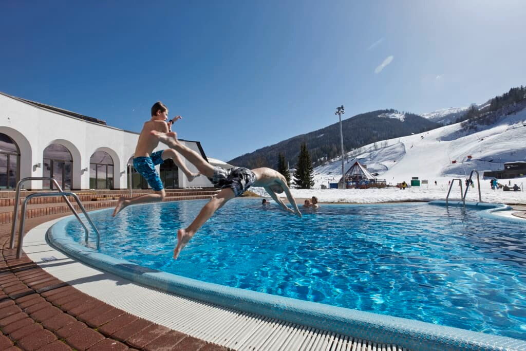 Zwei Personen springen in einen Swimmingpool mit schneebedeckten Bergen im Hintergrund