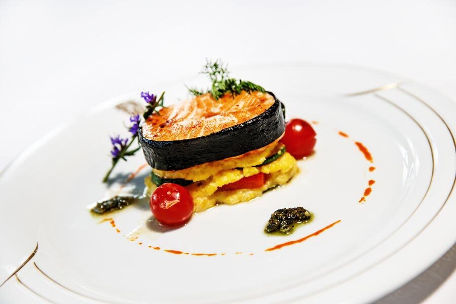 Ein Gourmetgericht mit einer Scheibe Lachs auf einem bunten Gemüsebett mit Garnierungen.