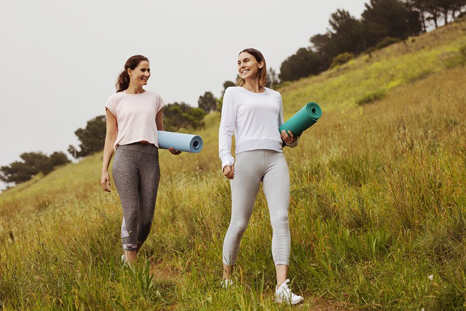 Zwei Frauen gehen mit Yoga-Matten über eine Wiese in der Natur zu einem Outdoor-Fitnessbereich.
