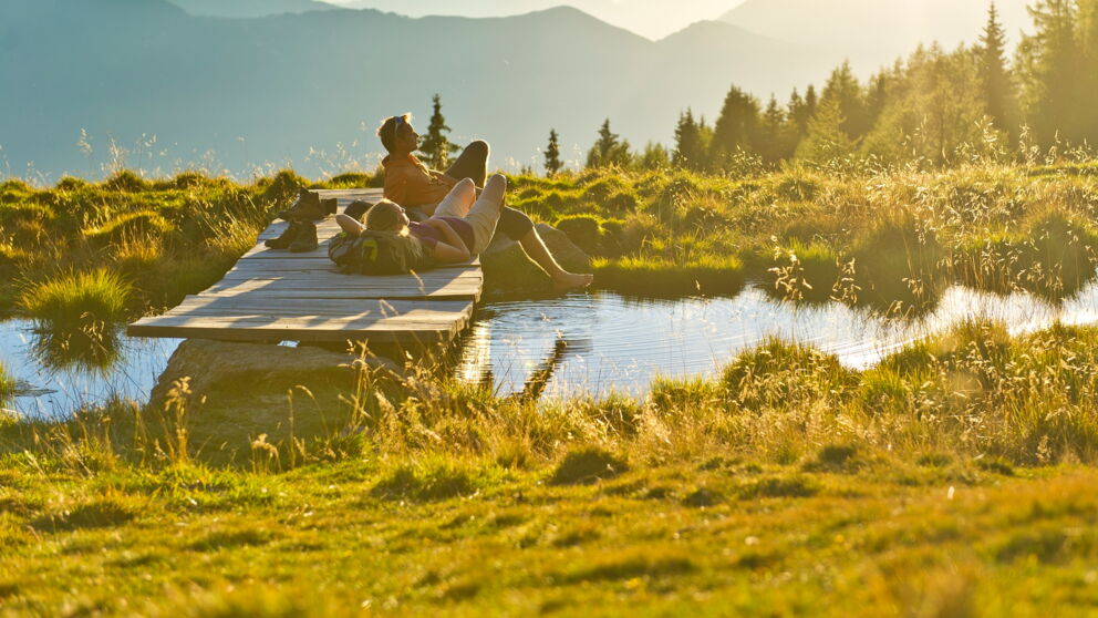 Two hikers lying on a small wooden bridge by a pond