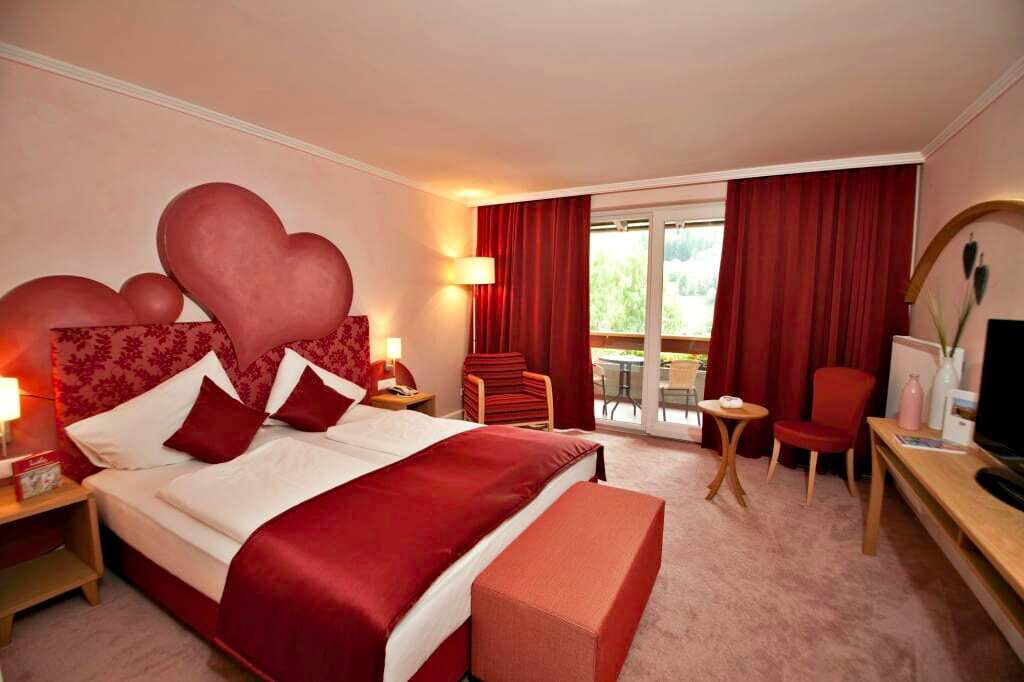 Elegantes Hotelzimmer mit Doppelbett und herzförmigem Kopfteil, warmes Licht, Balkonansicht