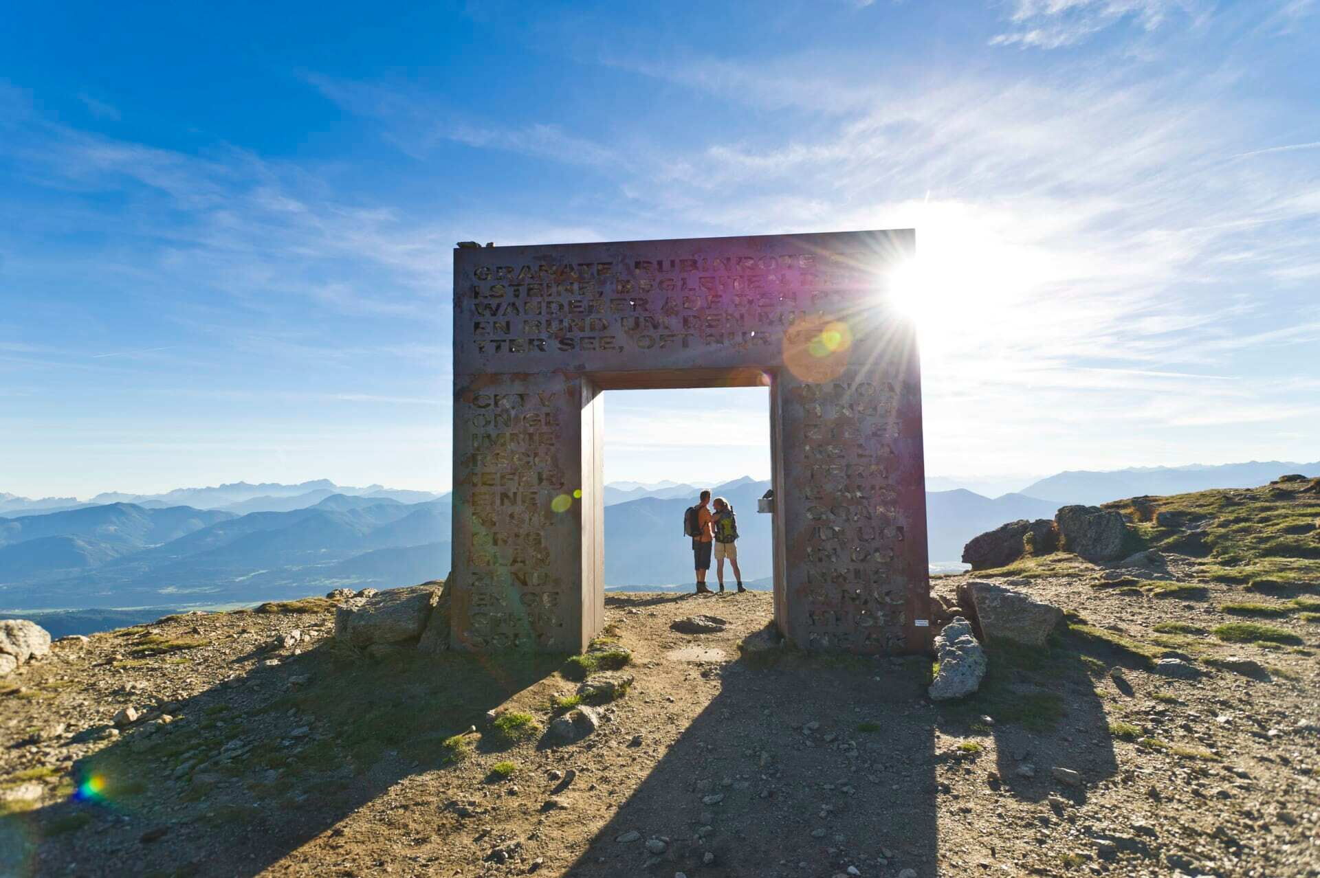 Zwei Wanderer stehen vor einem großen Steinmonument mit Panoramablick auf die Berglandschaft.